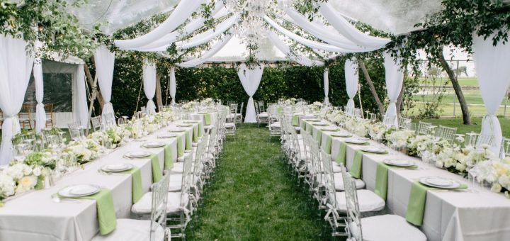 The dining area for an outdoor wedding with a large tent, hanging fabric, crystal chandeliers, and white chairs.