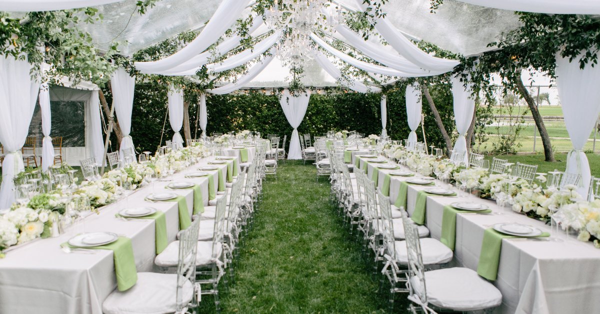 The dining area for an outdoor wedding with a large tent, hanging fabric, crystal chandeliers, and white chairs.