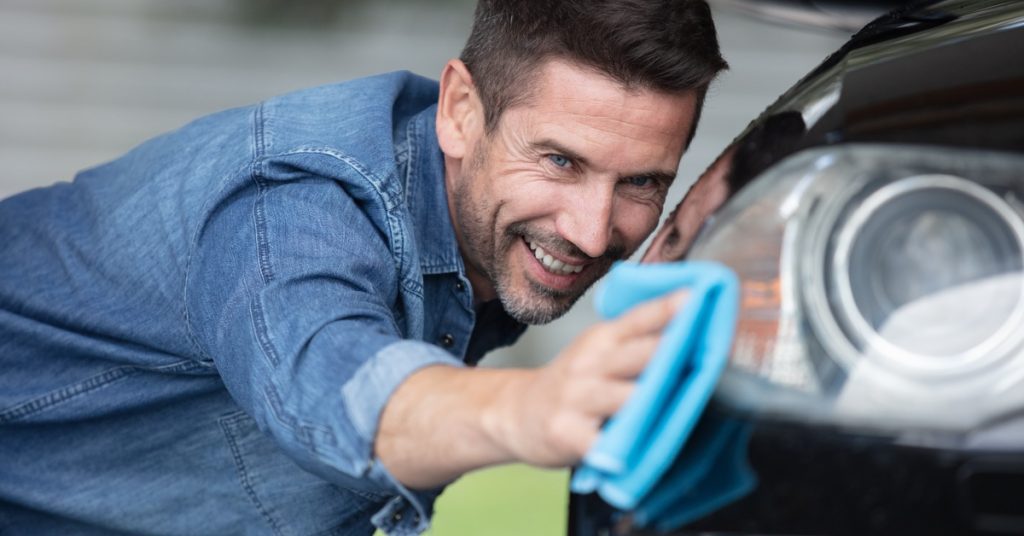 A smiling man cleaning the exterior of his car. He is running a microfiber cloth along the vehicle's headlight.