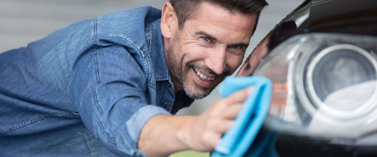 A smiling man cleaning the exterior of his car. He is running a microfiber cloth along the vehicle's headlight.