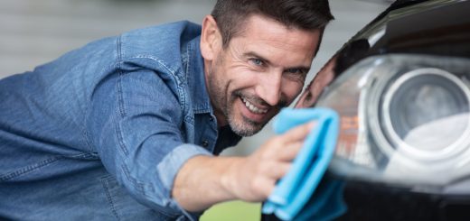 A smiling man cleaning the exterior of his car. He is running a microfiber cloth along the vehicle's headlight.