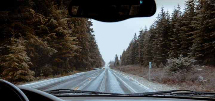 A person is driving down a road. The road is covered in snow and there are trees on either side of the road.