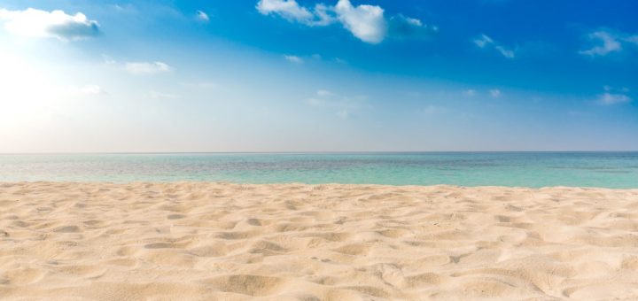 A wide sandy beach with soft ripples, calm turquoise ocean, and bright blue sky with scattered clouds on a sunny day.