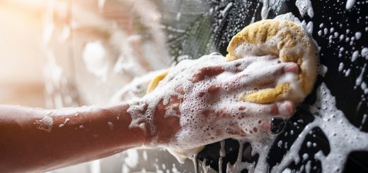 A close-up of someone's hand, circling a yellow sponge over a soapy black car to clean it. The sun reflects on the car.