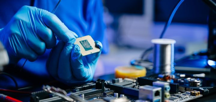 An engineer sits at their workstation as they wear blue latex gloves to hold and work on a microchip for an electronic.