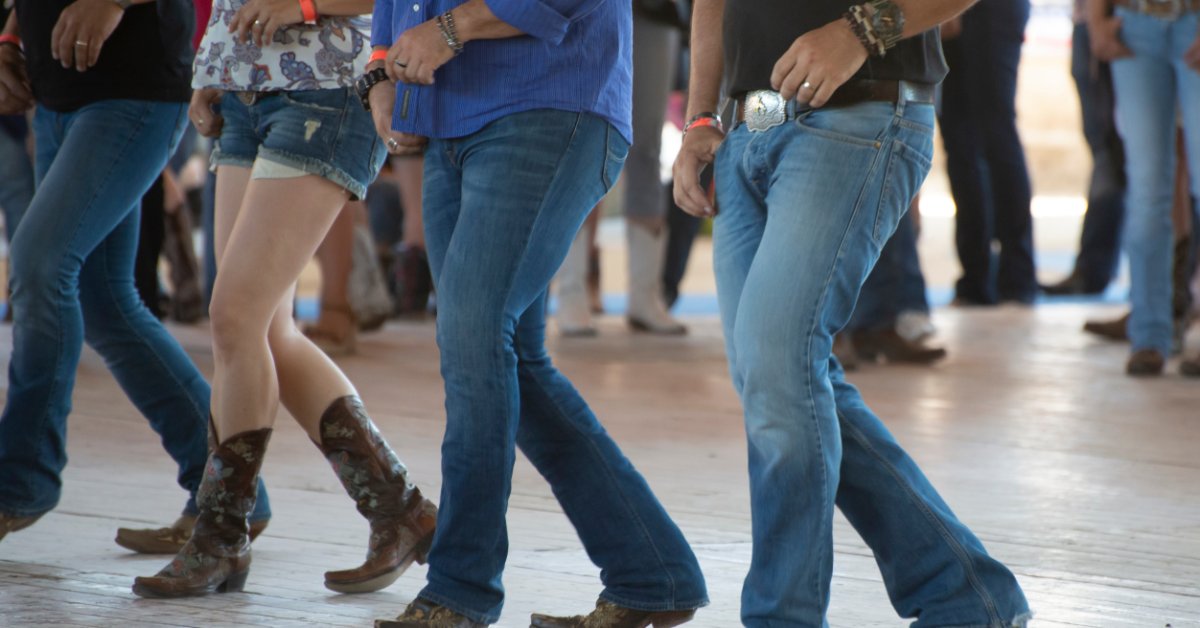 A group of adults wearing cowboy hats and boots line dancing together on a wooden dance floor indoors.