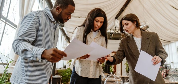 Three people holding paper documents in an event venue. The group looks at one of the documents together.