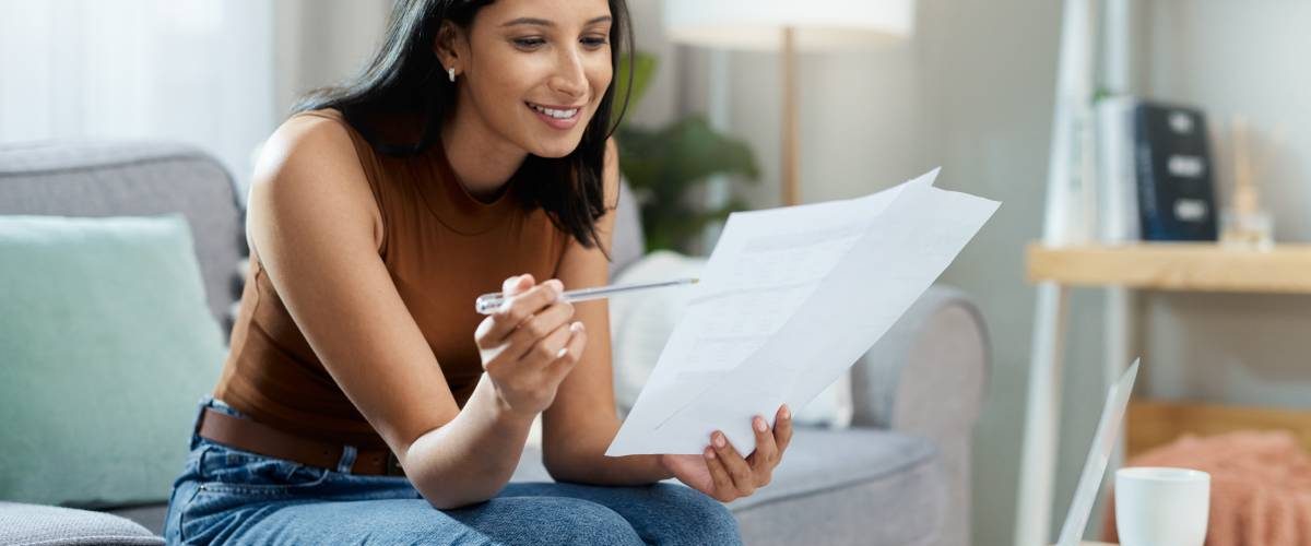A woman smiles as she sits at home, reviewing various documents with a pen. A laptop sits in front of her.