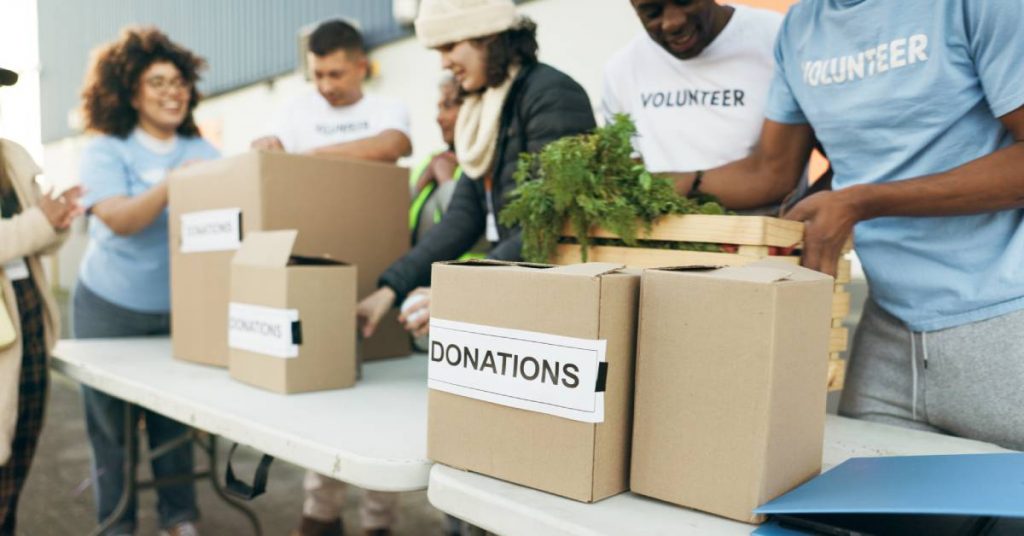 People standing behind a table, wearing shirts that say "volunteer." Boxes line the table labeled "donations."