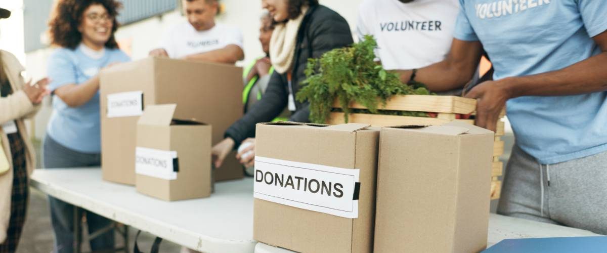 People standing behind a table, wearing shirts that say "volunteer." Boxes line the table labeled "donations."