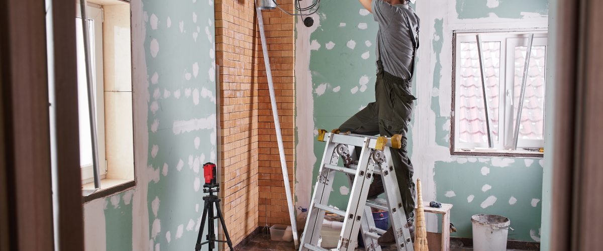 A contractor stands on a ladder as they work on the ceiling in a room. The room has been gutted to the studs.