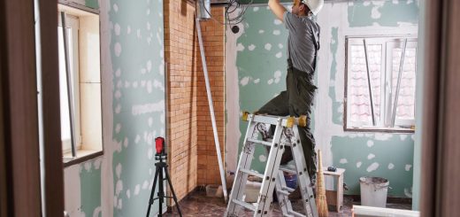 A contractor stands on a ladder as they work on the ceiling in a room. The room has been gutted to the studs.