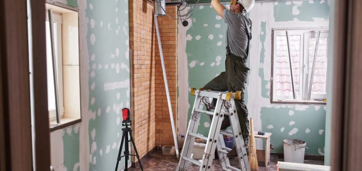 A contractor stands on a ladder as they work on the ceiling in a room. The room has been gutted to the studs.