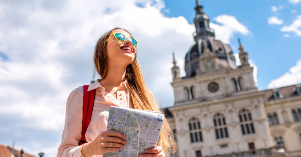 A young female travels alone. She holds a map and wears sunglasses and a red backpack in the city center.