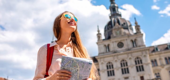 A young female travels alone. She holds a map and wears sunglasses and a red backpack in the city center.