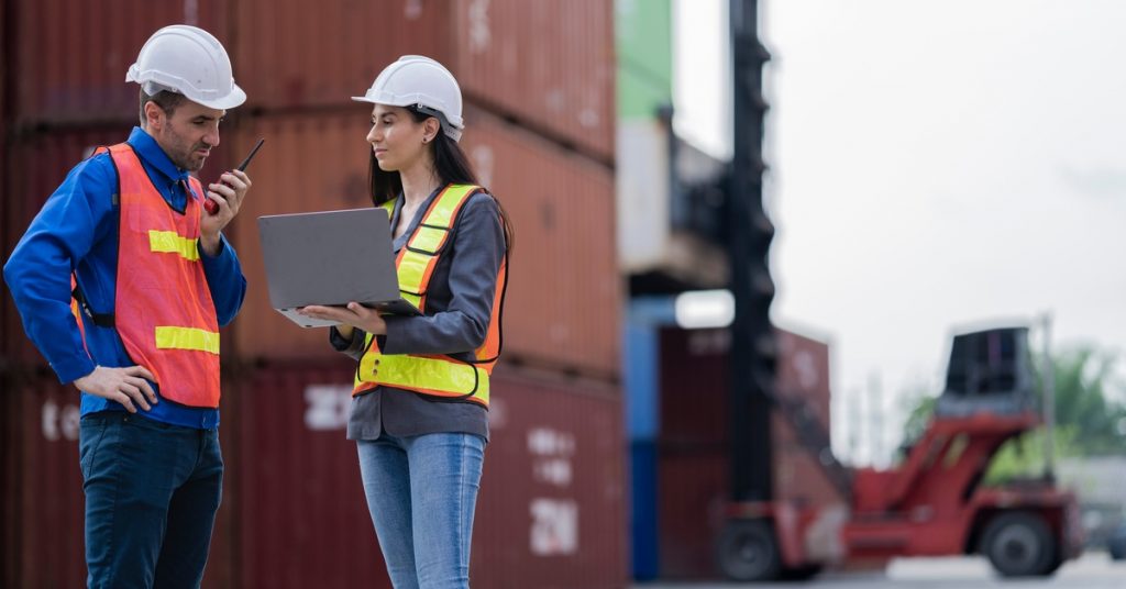 Two logistics workers in safety vests and hard hats standing in front of a stack of shipping containers.