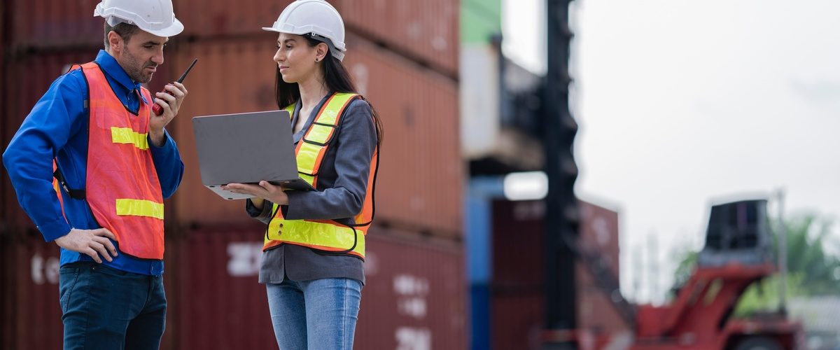 Two logistics workers in safety vests and hard hats standing in front of a stack of shipping containers.