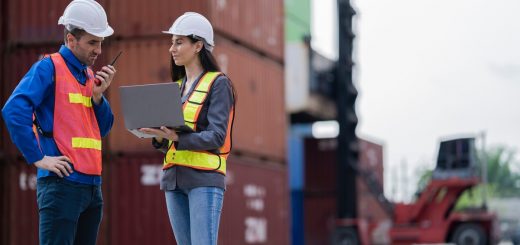 Two logistics workers in safety vests and hard hats standing in front of a stack of shipping containers.