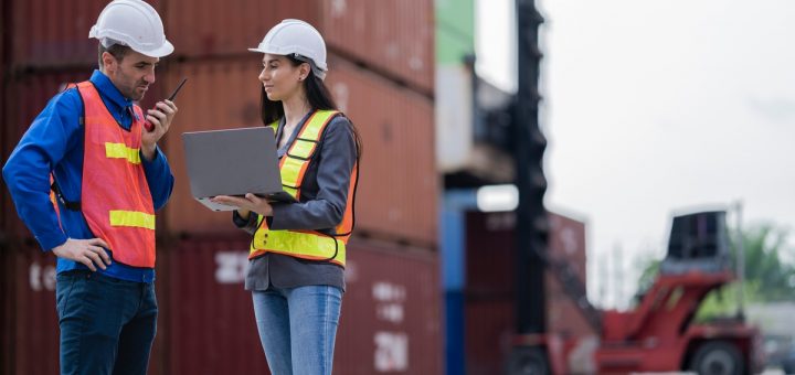 Two logistics workers in safety vests and hard hats standing in front of a stack of shipping containers.