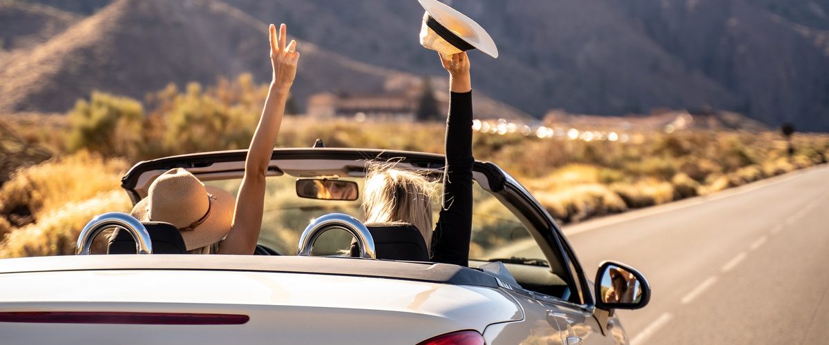 Two women with hats driving down an open road in the desert. They both have one hand up, celebrating.