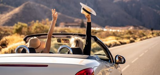 Two women with hats driving down an open road in the desert. They both have one hand up, celebrating.