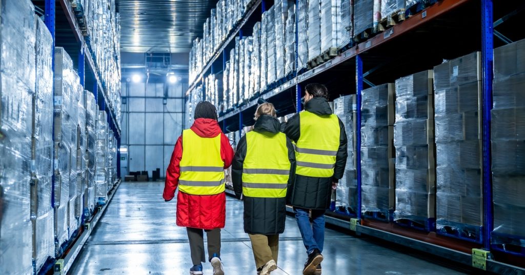 Three team members wearing heavy jackets under neon-yellow safety vests are walking in a warehouse.