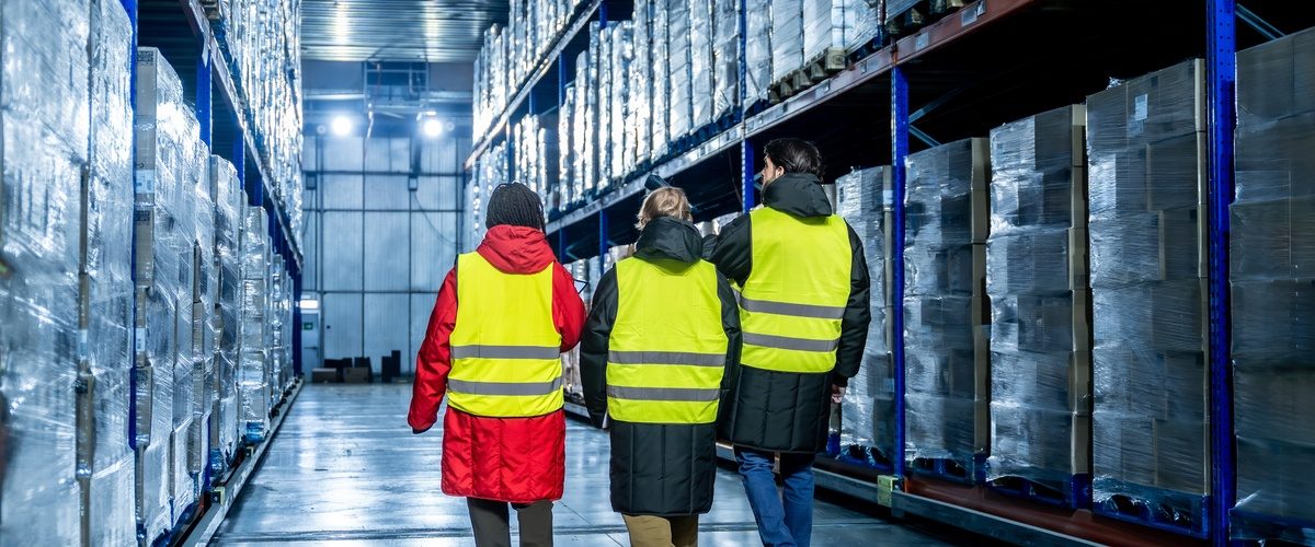 Three team members wearing heavy jackets under neon-yellow safety vests are walking in a warehouse.