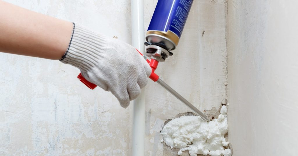A person is holding a spray foam gun and is using it to install foam into a crack on a concrete wall.