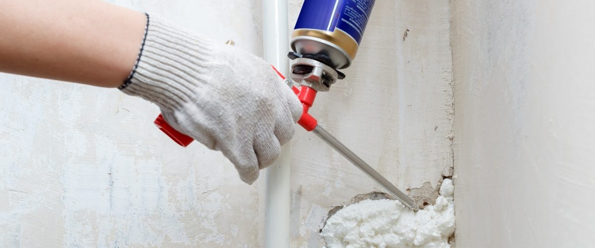 A person is holding a spray foam gun and is using it to install foam into a crack on a concrete wall.