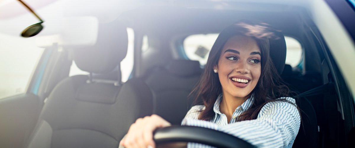 A happy woman driving in a car and smiling. She has one hand on the wheel and is looking out the driver's window.