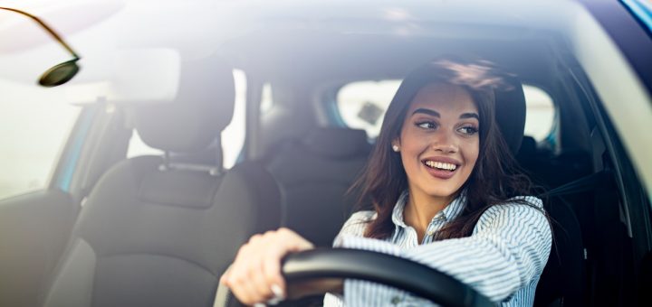 A happy woman driving in a car and smiling. She has one hand on the wheel and is looking out the driver's window.