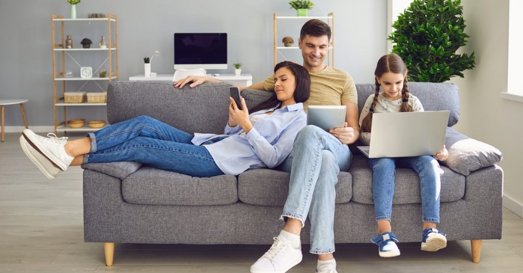 A family of three shares their gray couch in the living room as each person is on a tech device using the internet.