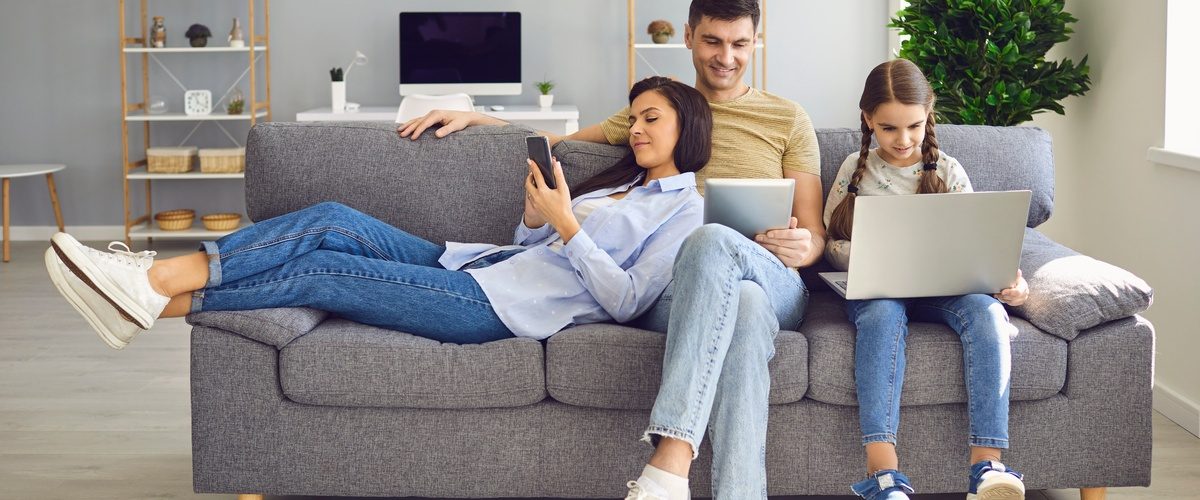 A family of three shares their gray couch in the living room as each person is on a tech device using the internet.