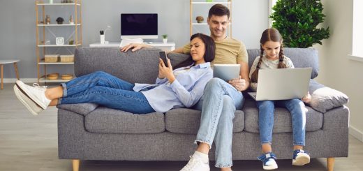 A family of three shares their gray couch in the living room as each person is on a tech device using the internet.