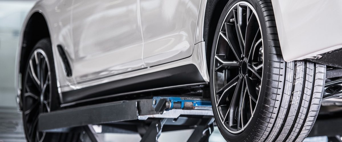 A white vehicle lifted in a repair shop with the driver's side tires exposed. The tires appear brand-new.