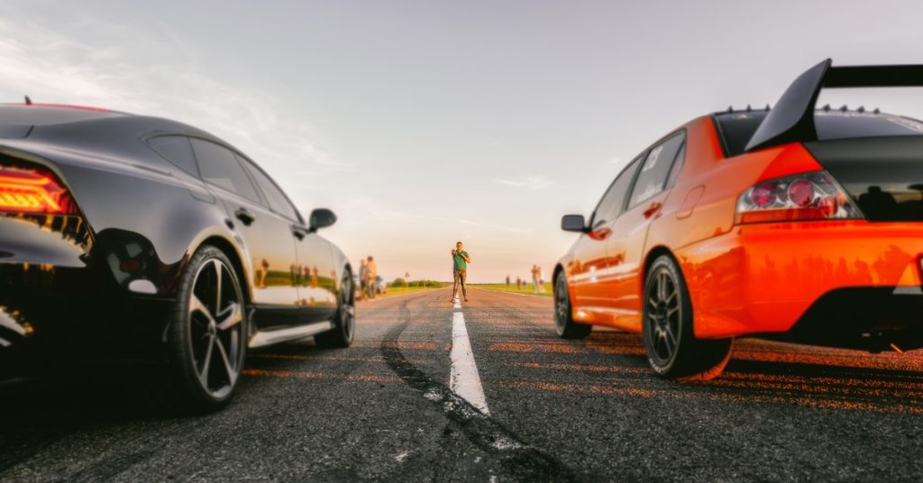 Two sports cars are side by side on a long stretch of road. A person in a green shirt stands between them at a distance.