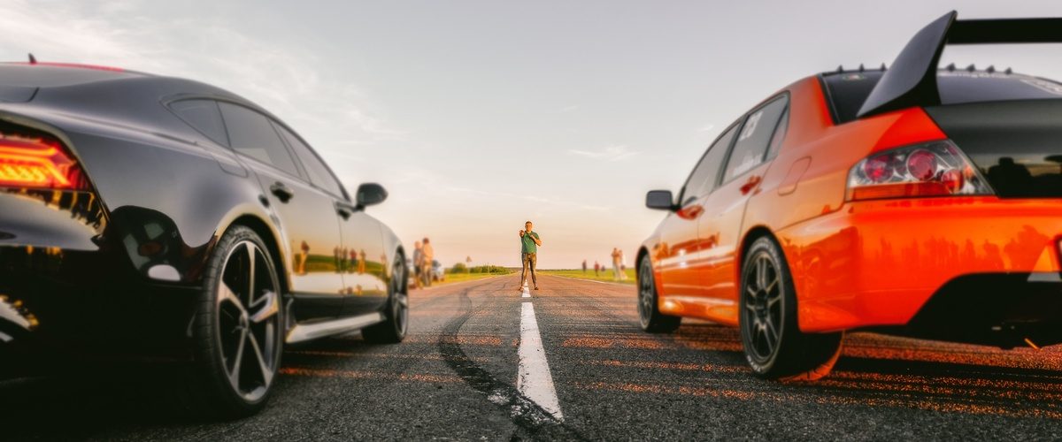 Two sports cars are side by side on a long stretch of road. A person in a green shirt stands between them at a distance.