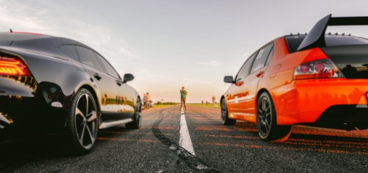 Two sports cars are side by side on a long stretch of road. A person in a green shirt stands between them at a distance.