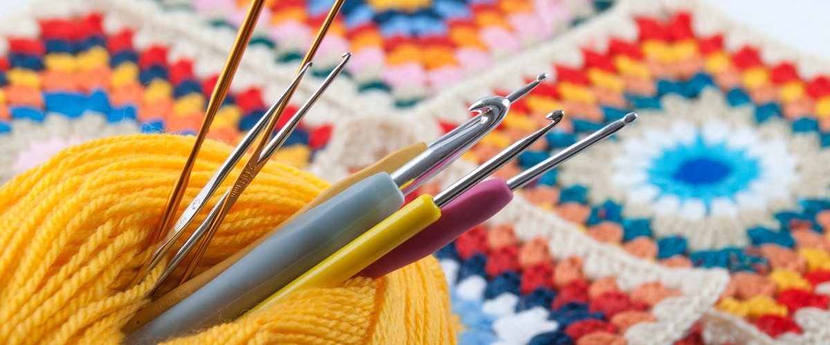 Several silver crochet hooks are inserted through a skein of yellow thread resting on a blue knitted surface.