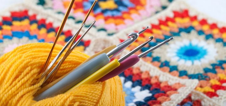 Several silver crochet hooks are inserted through a skein of yellow thread resting on a blue knitted surface.
