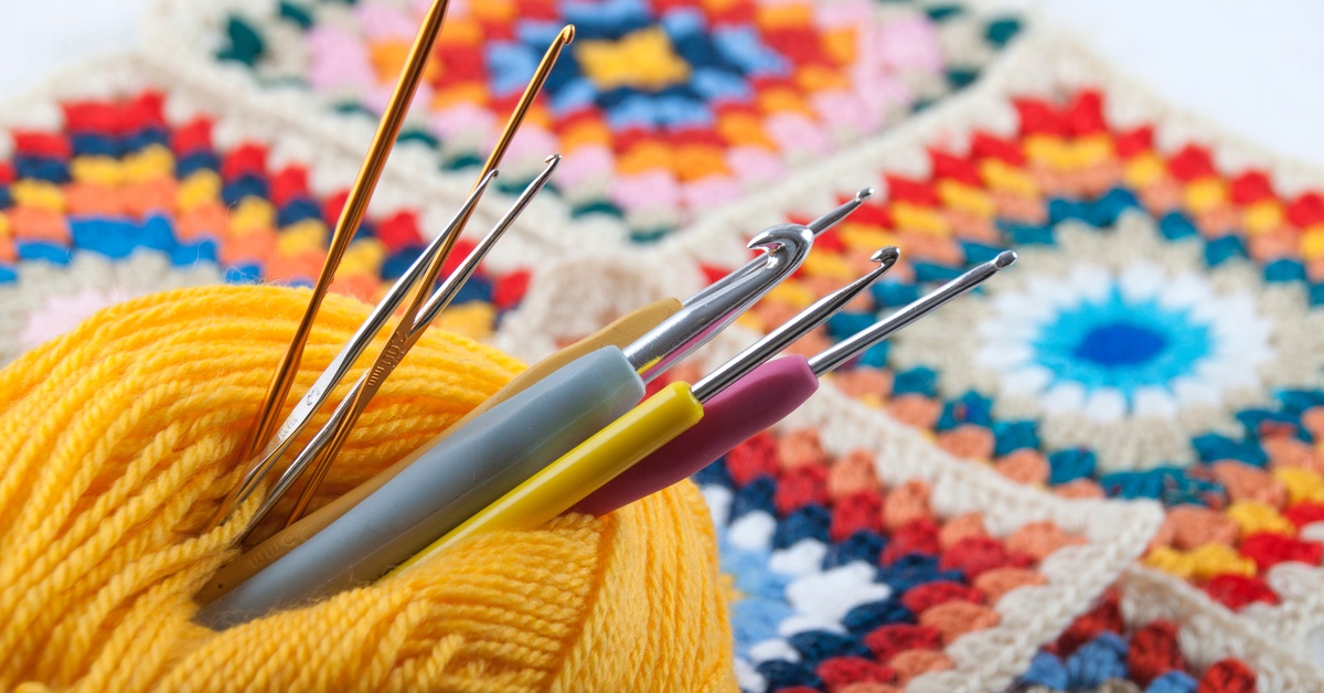 Several silver crochet hooks are inserted through a skein of yellow thread resting on a blue knitted surface.
