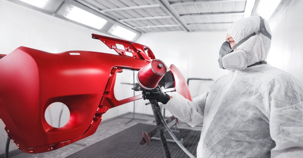 Worker in protective suit and respirator spray-paints a glossy red car bumper inside an automotive paint booth.