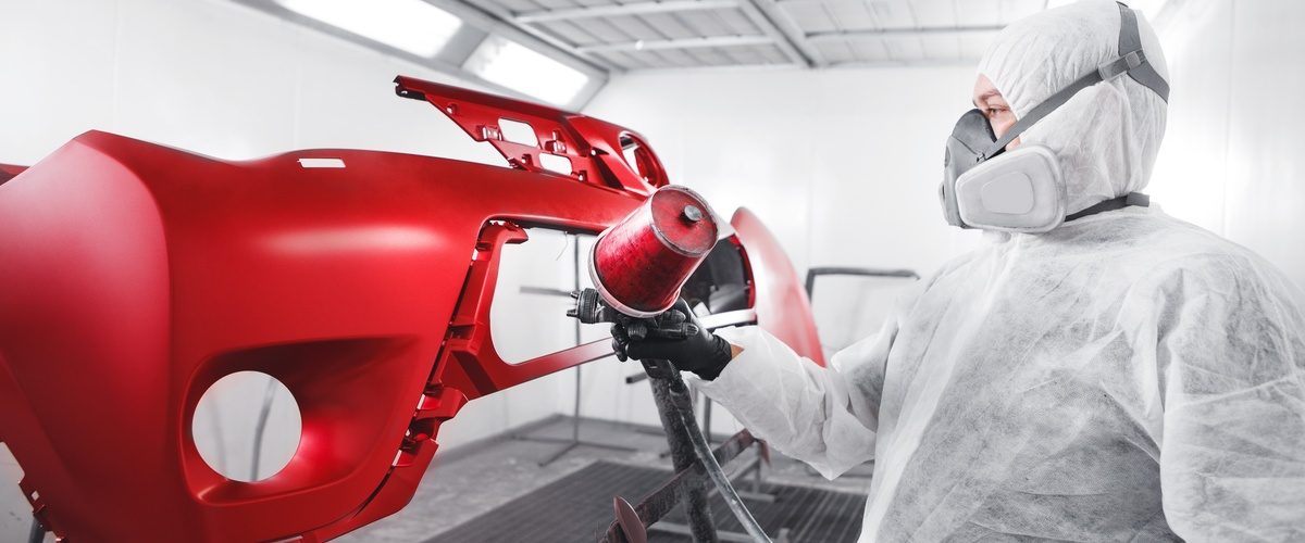 Worker in protective suit and respirator spray-paints a glossy red car bumper inside an automotive paint booth.