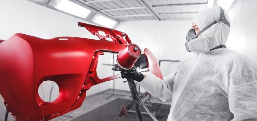 Worker in protective suit and respirator spray-paints a glossy red car bumper inside an automotive paint booth.