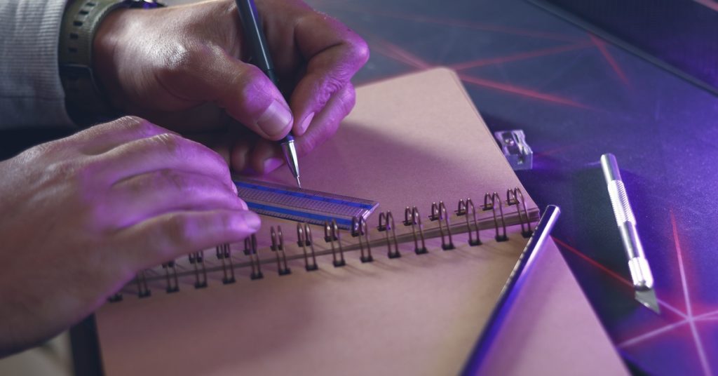 A left-handed person drawing a line with a pen and ruler in a spiral sketchbook with purple lighting and a cutting tool nearby.
