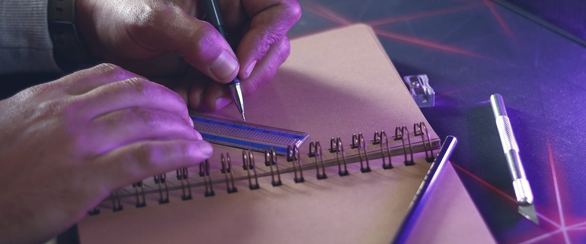 A left-handed person drawing a line with a pen and ruler in a spiral sketchbook with purple lighting and a cutting tool nearby.