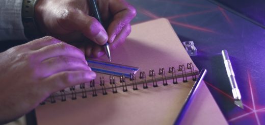 A left-handed person drawing a line with a pen and ruler in a spiral sketchbook with purple lighting and a cutting tool nearby.