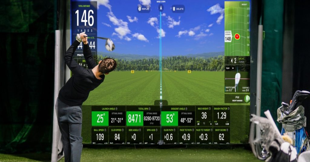 A golfer swinging a club indoors toward a digital golf screen with yardage metrics, green turf, and golf bags nearby.