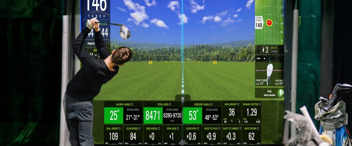 A golfer swinging a club indoors toward a digital golf screen with yardage metrics, green turf, and golf bags nearby.