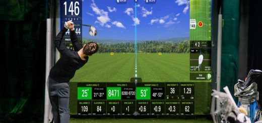 A golfer swinging a club indoors toward a digital golf screen with yardage metrics, green turf, and golf bags nearby.